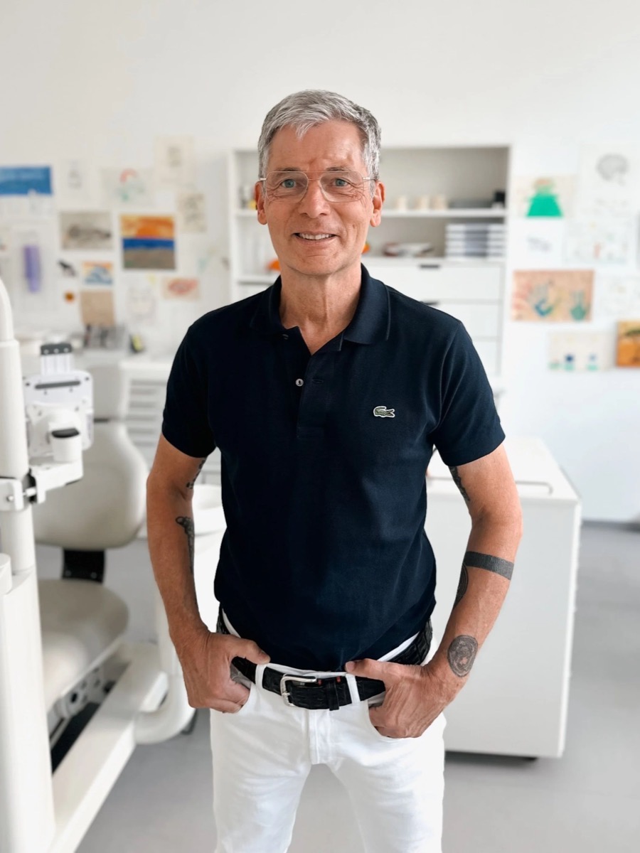 Dr. Michael Baron – ein älterer Mann mit Brille und grauem Haar steht lachend in einem Zimmer mit Arzt- oder Zahnarzt-Ausstattung, trägt ein dunkles Lacoste-Poloshirt und weiße Hosen, und hat Tätowierungen am Arm.