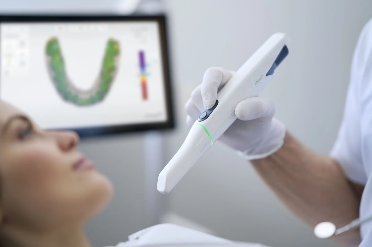 Ein Zahnarzt hält ein digitales Diagnostikgerät über den Mund einer Patientin im Behandlungszimmer. Im Hintergrund ein Bildschirm mit einer digitalen Abbildung des Zahn- und Kieferbereichs.