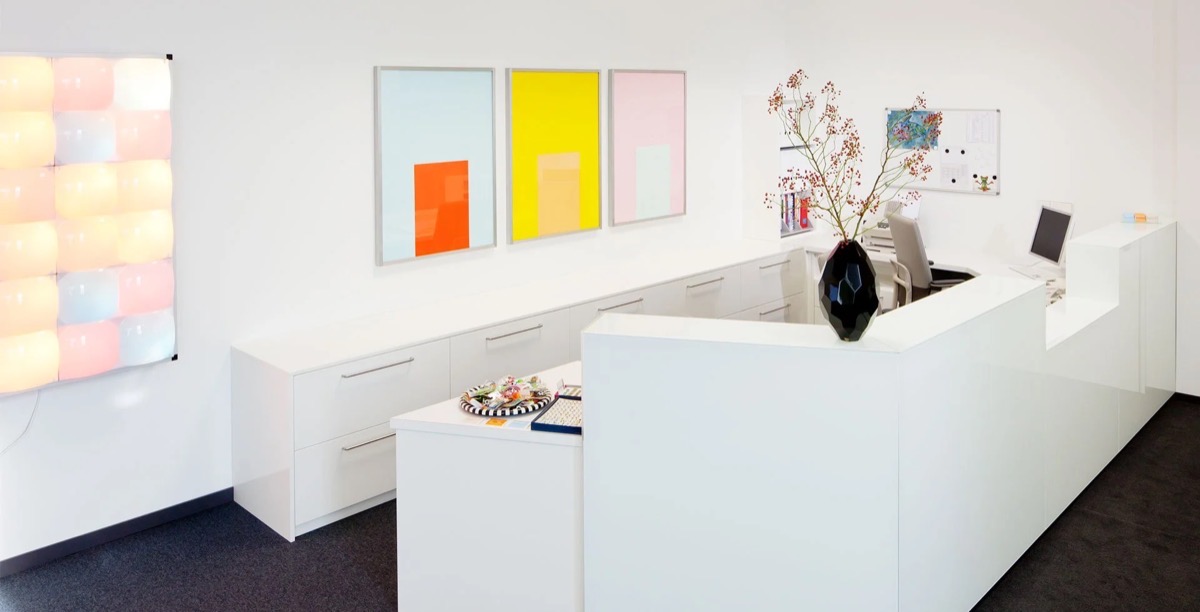 Helle Arztpraxis mit weißem Empfangstresen, Wandkunst in Pastellfarben, Vase mit Zweigen auf dem Empfangstresen, Computer und Akten auf dem Schreibtisch, dunkler Teppichboden.