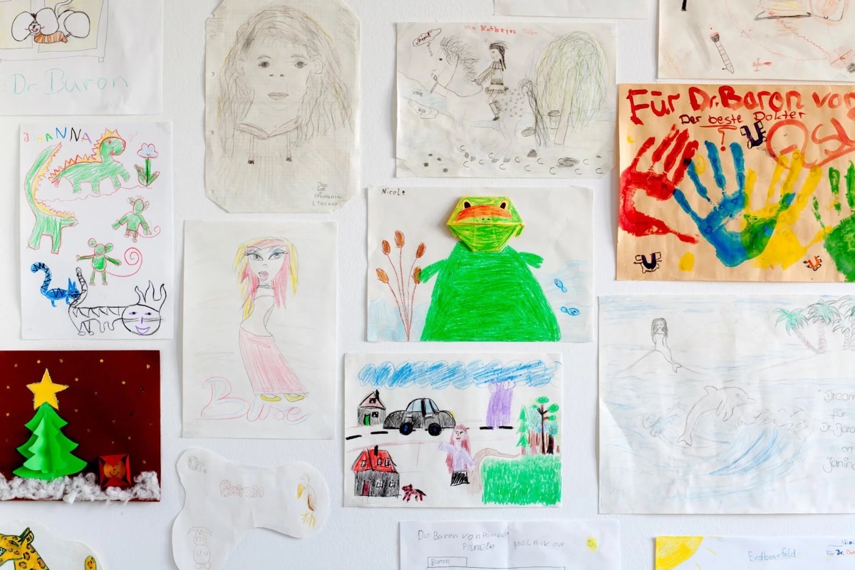 Verschiedene Kinderzeichnungen und Gemälde, darunter eine farbige Zeichnung eines Frosches, eine Silhouette einer Frau, ein bunter Dinosaurier sowie ein Weihnachtsbaum, alles an einer weißen Wand befestigt.