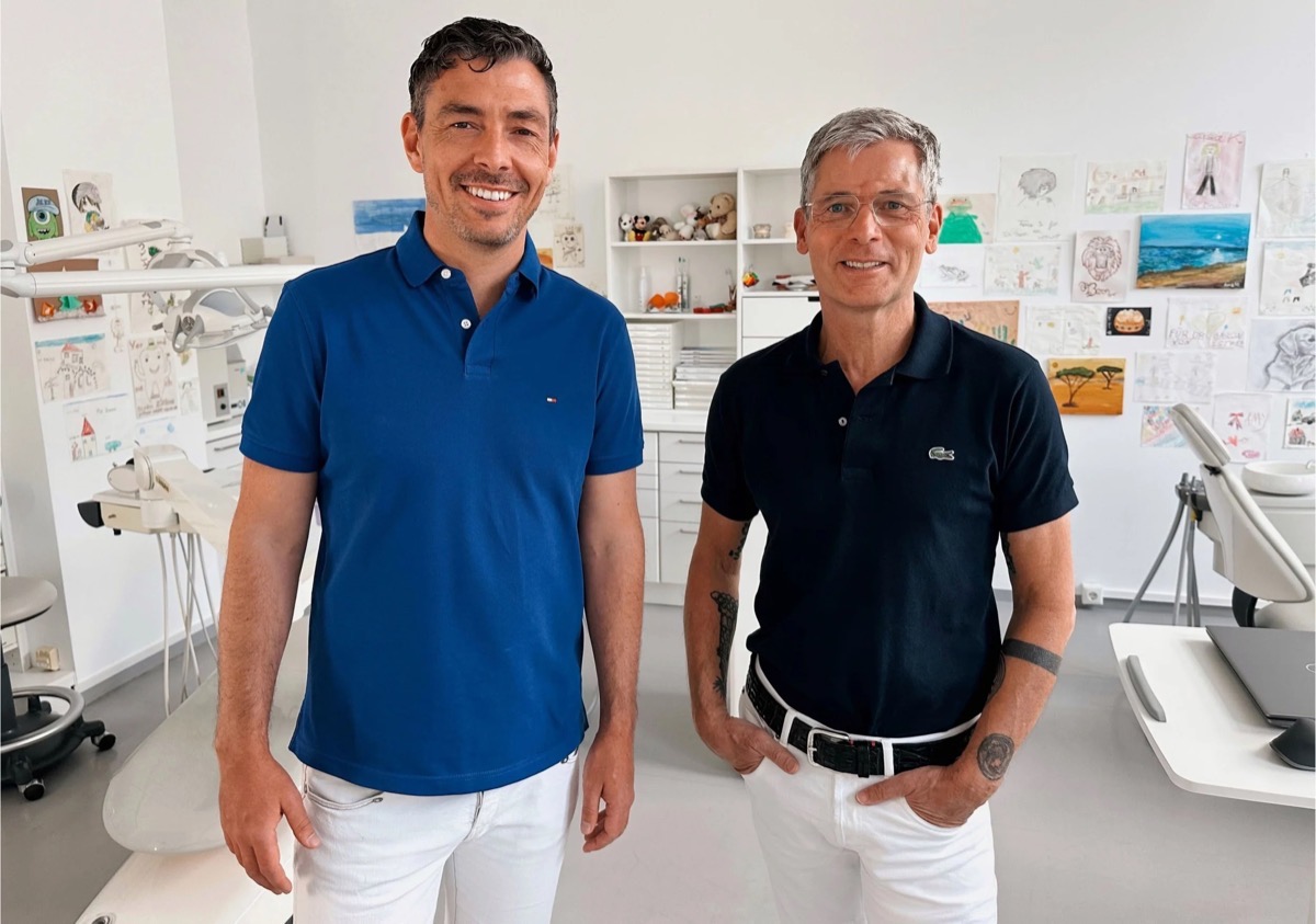 Zwei Männer stehen in einer Arztpraxis, beide tragen schwarze Poloshirts und weiße Hosen, lächeln und blicken in die Kamera. Dr. Christoph Dippold und Dr. Michael Baron sind das Ärzteteam der Kieferorthopädie an den Erlanger Höfen.